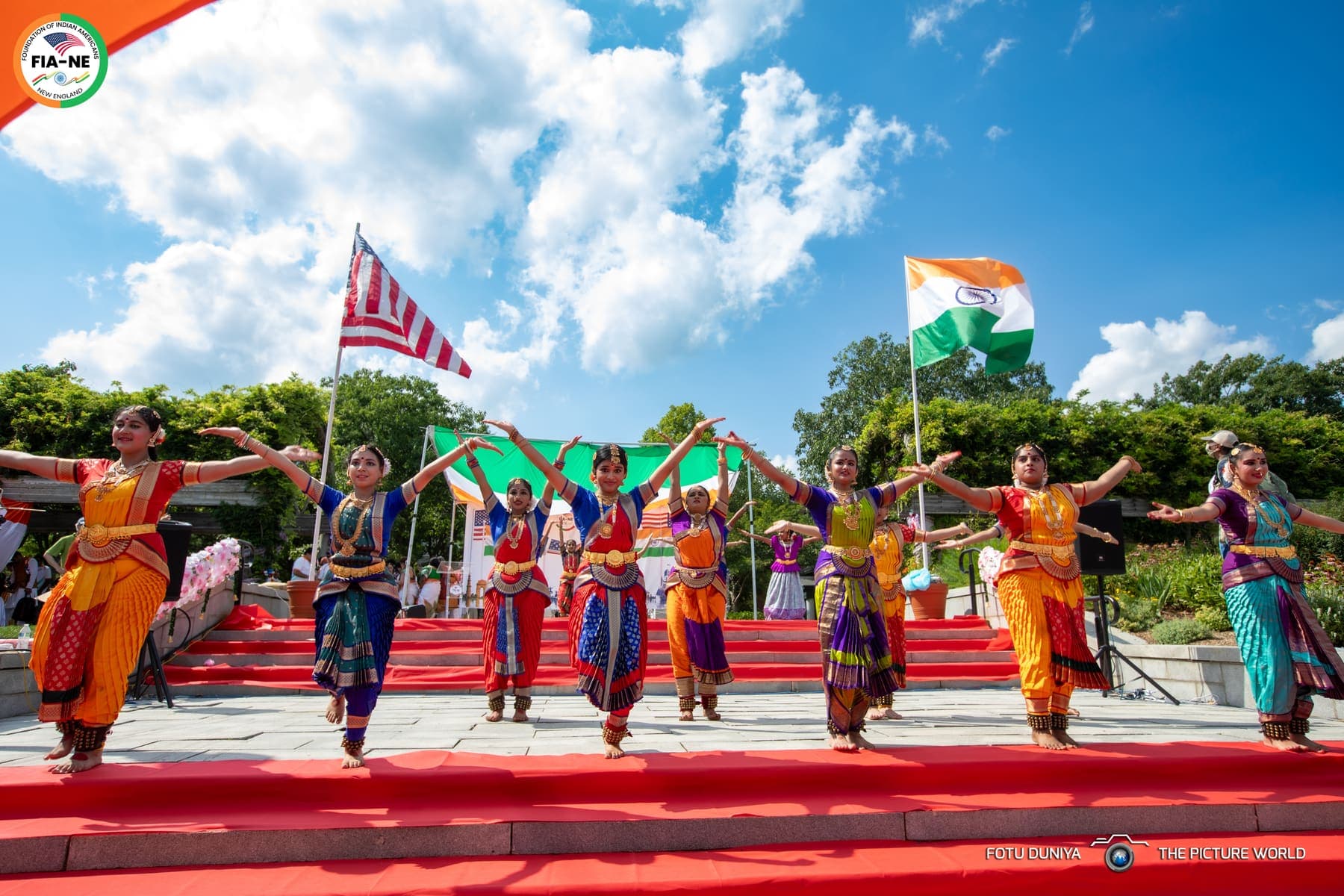 A classical Indian dance troupe performing on an outdoor stage with Indian and American flags flying behind them
