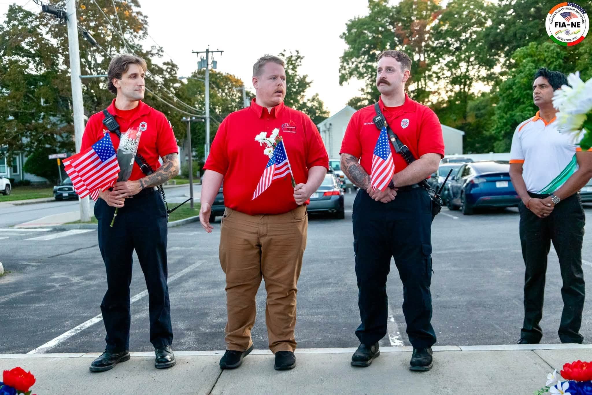 Three Westborough firefighters in red shirts standing at attention holding small American flags