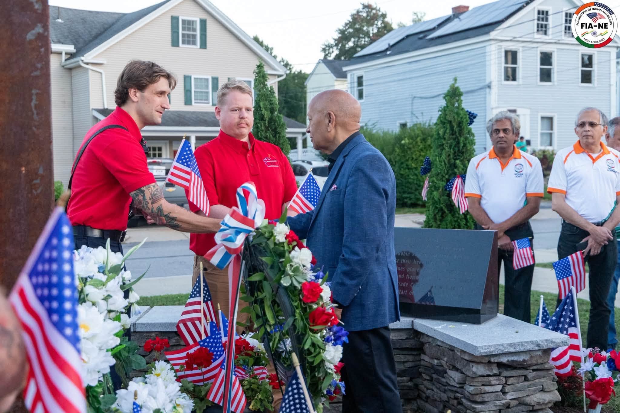 A firefighter handing a small American flag to a community elder beside the memorial plaque