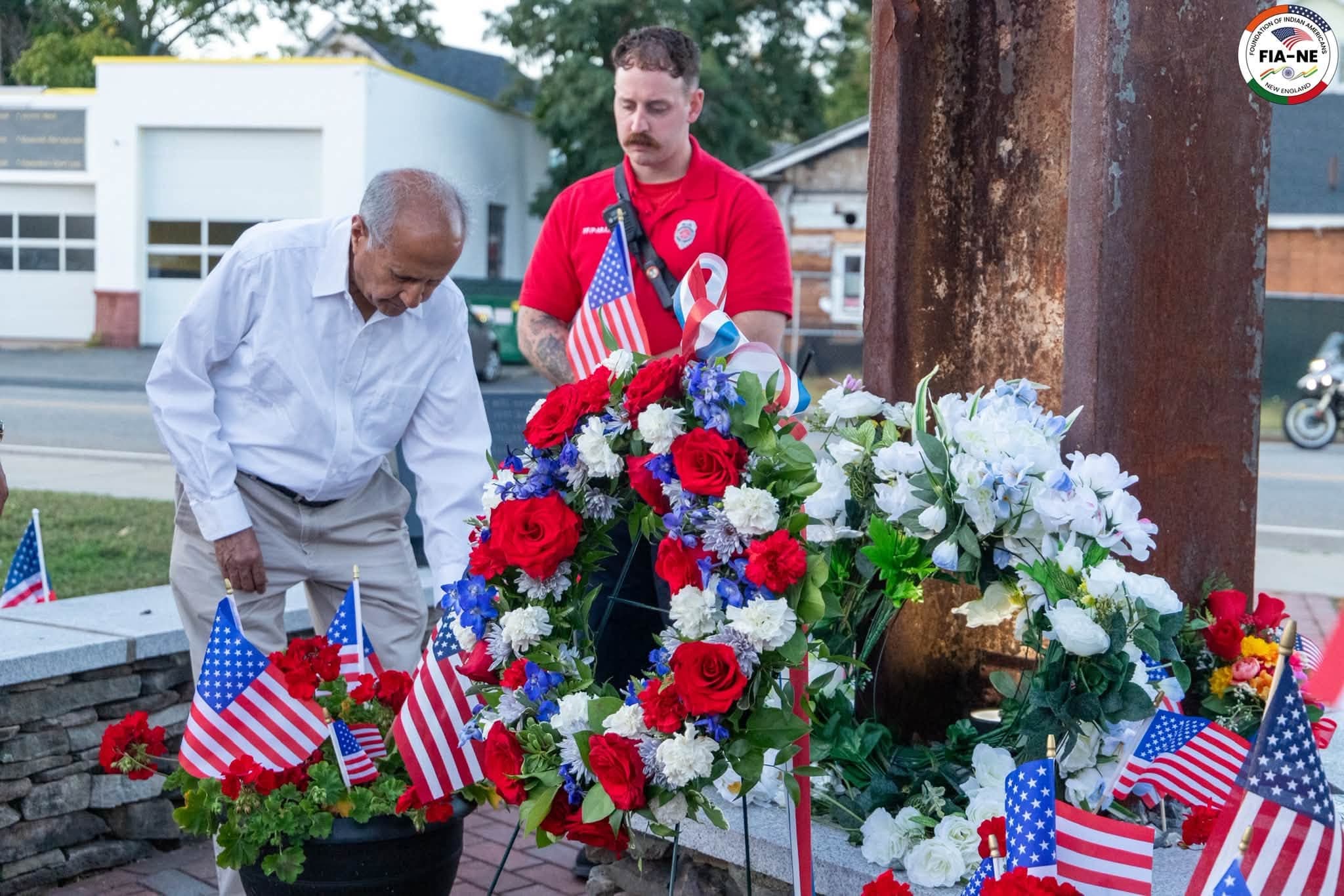 A firefighter and a FIANE member placing a wreath of red, white, and blue flowers at the base of the steel-beam memorial